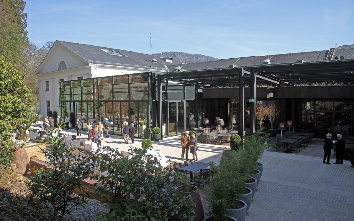 Terrasse mit dunklen Café-Möbeln und einem Glaspavillon neben dem Casino-Flügel des Kurhauses, Besucher in der Sonne