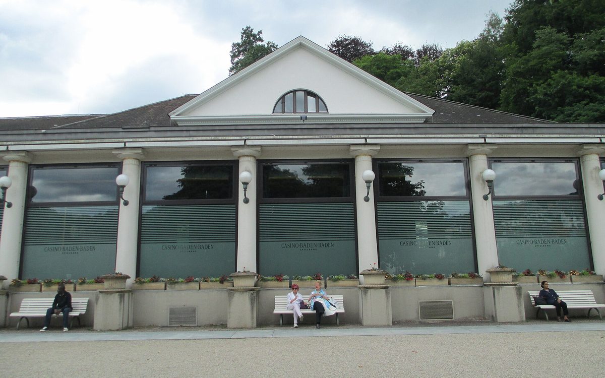 Casino-Flügel des Kurhauses mit Giebel, weißen Säulen und Fenstern mit der Aufschrift Casino Baden-Baden, davor Besucher auf Bänken